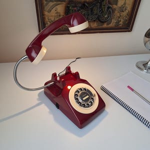 May include: A burgundy rotary phone-shaped desk lamp with a cream-colored handset. The lamp has a flexible metal neck and a circular dial. A chrome alarm clock and a notepad with a pen are also visible.