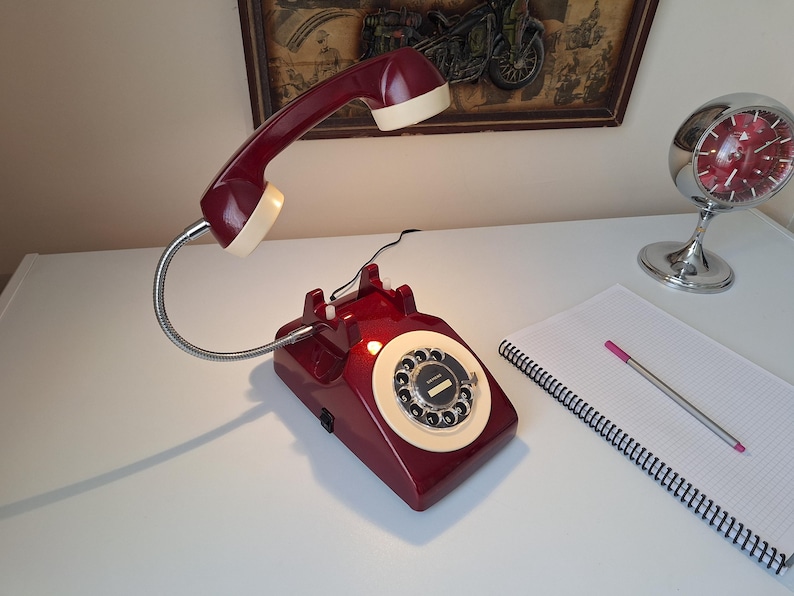 May include: A burgundy rotary phone-shaped desk lamp with a cream-colored handset. The lamp has a flexible metal neck and a circular dial. A chrome alarm clock and a notepad with a pen are also visible.