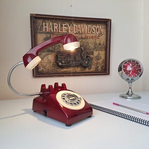 May include: A burgundy telephone-shaped desk lamp with a flexible chrome neck and a cream-colored receiver. The lamp is on a white surface, with a notepad, pencil, and a vintage Harley-Davidson sign in the background. A chrome clock is also visible.
