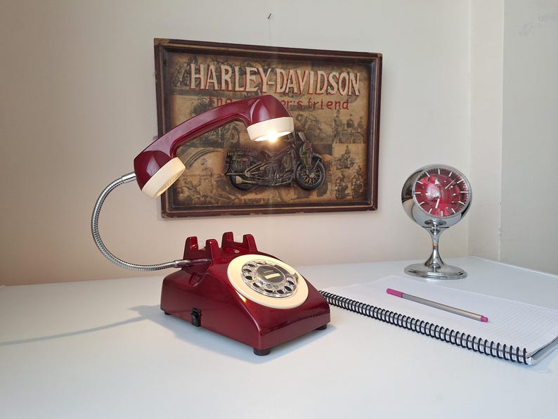 May include: A burgundy telephone-shaped desk lamp with a flexible chrome neck and a cream-colored receiver. The lamp is on a white surface, with a notepad, pencil, and a vintage Harley-Davidson sign in the background. A chrome clock is also visible.