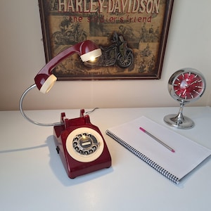 May include: A burgundy rotary phone lamp with a white dial and handset, and a flexible chrome neck. A vintage-style clock and a notepad with a pen are also on the white surface. A Harley-Davidson poster is in the background.