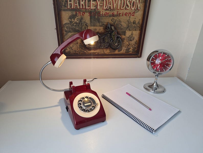May include: A burgundy rotary phone lamp with a white dial and handset, and a flexible chrome neck. A vintage-style clock and a notepad with a pen are also on the white surface. A Harley-Davidson poster is in the background.