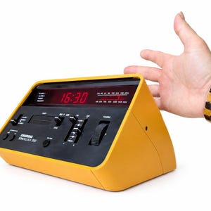 May include: A yellow Grundig Sonoclock 200 alarm clock radio with a black front panel. The device features a red digital display showing 16:30, a radio frequency tuner with AM and FM scales, sliding volume and tone controls, and push buttons.