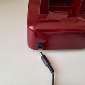 May include: A close-up of a burgundy-colored electronic device with a black power input port. A black power adapter cable is plugged into the device. The device is on a white surface.