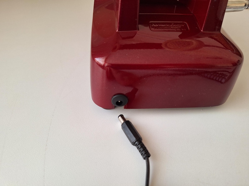 May include: A close-up of a burgundy-colored electronic device with a black power input port. A black power adapter cable is plugged into the device. The device is on a white surface.