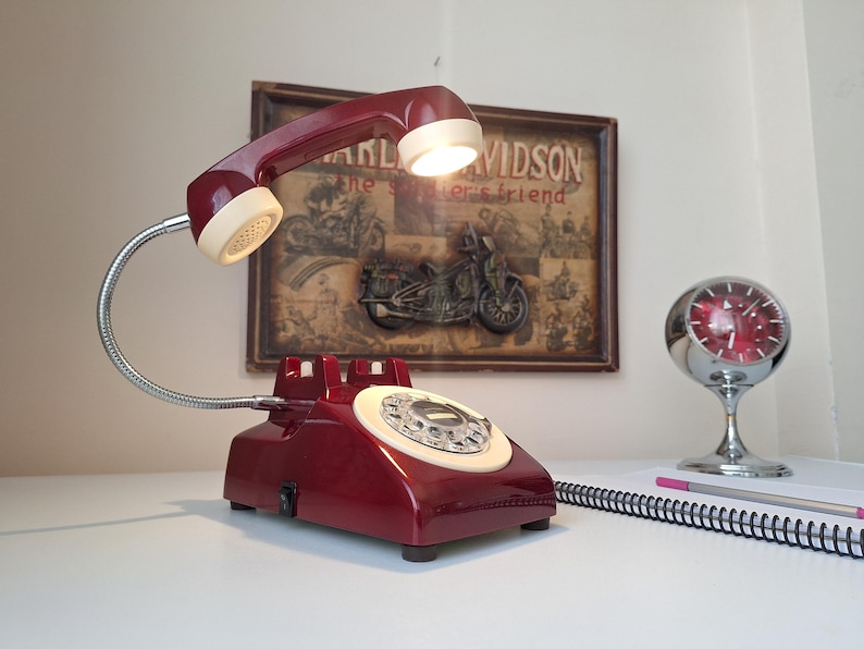 May include: A vintage-style desk lamp shaped like a rotary phone, in a deep red color with a cream-colored dial and handset. The lamp has a flexible, silver-colored gooseneck and a small lightbulb. A framed Harley-Davidson poster is in the background.