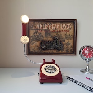 May include: A vintage burgundy rotary phone with a cream-colored dial and handset. The phone has a flexible silver cord and a small light on the handset. A Harley-Davidson sign and a chrome clock are in the background.