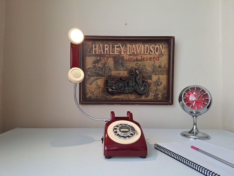 May include: A vintage burgundy rotary phone with a cream-colored dial and handset. The phone has a flexible silver cord and a small light on the handset. A Harley-Davidson sign and a chrome clock are in the background.