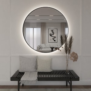 Puede incluir: Un banco negro con una manta blanca y dos almohadas blancas se encuentra frente a una pared blanca con un espejo redondo negro con una luz blanca alrededor del borde. El espejo refleja una habitación con un sofá blanco y una mesa de café negra.