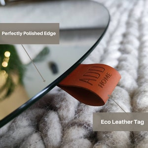 May include: A close-up of a round mirror with a perfectly polished edge. An eco-leather tag with the words "ADD HOME" is attached to the mirror. The mirror is resting on a textured, off-white surface.