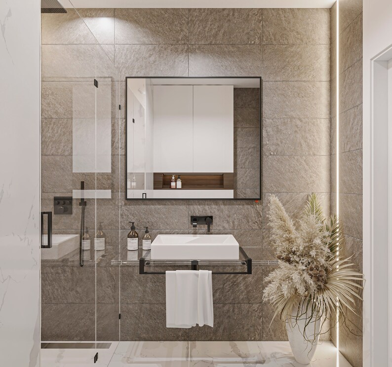 Puede incluir: Un ba&ntilde;o moderno con un lavabo de porcelana blanca, un espejo con marco negro y una cabina de ducha de vidrio. Las paredes est&aacute;n revestidas con azulejos de color marr&oacute;n claro y una toalla blanca cuelga de un toallero negro.