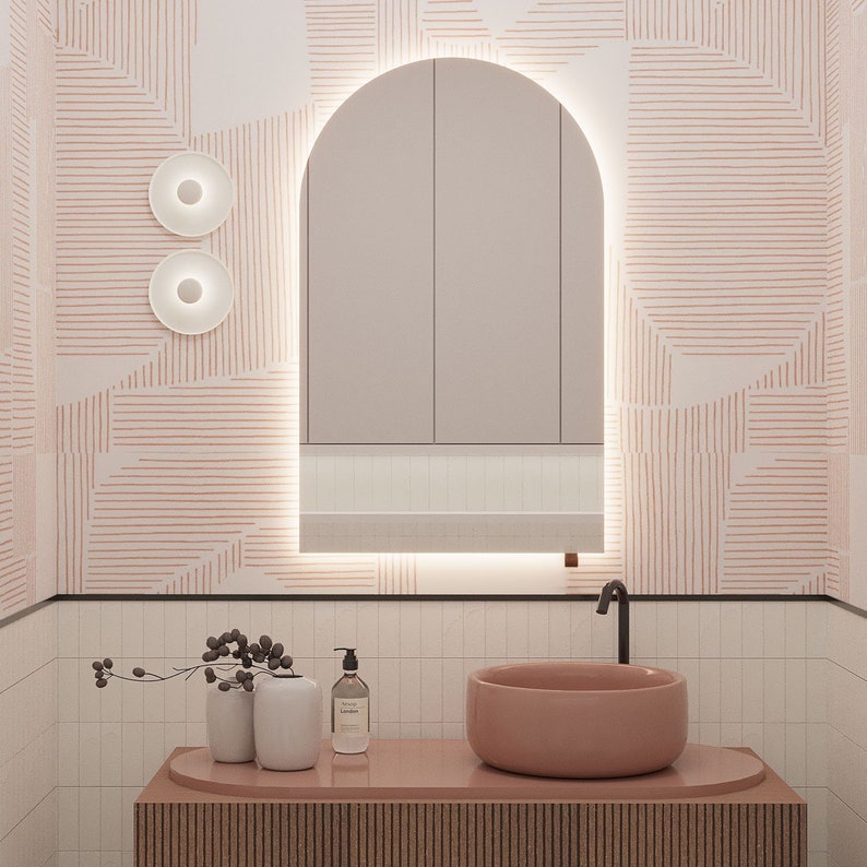 May include: A modern bathroom with a pink color scheme. The room features a large arched mirror with integrated lighting, a pink vessel sink, and a wooden vanity. The walls are decorated with a geometric pattern in pink and white, and two round wall sconces are visible.
