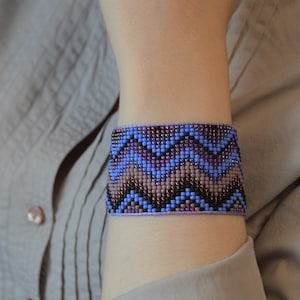 May include: A beaded bracelet with a geometric pattern in shades of blue, purple, and brown. The bracelet is made with small, colorful beads and has a woven design.