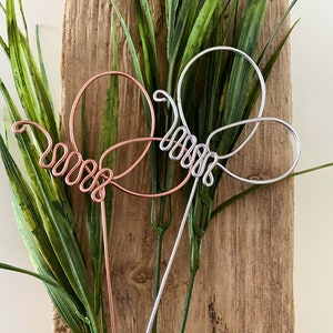 May include: Two wire butterfly garden stakes, one silver and one copper, against a wood background with green foliage.