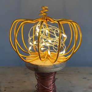 May include: A gold wire crown with a string of white fairy lights inside. The crown is sitting on a wooden spool.