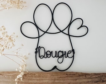 以下が含まれることがあります： 黒いワイヤー製の足跡ウォールアート。名前「Dougie」が筆記体で書かれています。足跡のデザインは、2つの大きなループと2つのハート型の要素が特徴です。アートは白い背景と流木の上に展示されています。