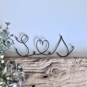 May include: Black wire sculpture of the word "Seas" with a heart in place of the "a". The sculpture is sitting on a piece of weathered wood.