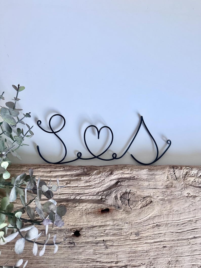 May include: Black wire sculpture of the word "Seas" with a heart in place of the "a". The sculpture is sitting on a piece of weathered wood.