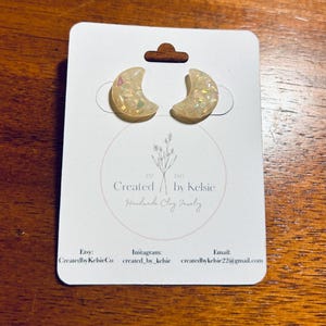 May include: A pair of white crescent moon earrings with iridescent glitter embedded in the clay. The earrings are displayed on a white card with the text "Created by Kelsie Handmade Clay Jewelry" and contact information.