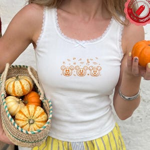 May include: A white ribbed tank top with a lace-trimmed scoop neckline and a small bow at the center. The chest features an orange graphic print of three pumpkin-shaped mouse heads surrounded by small bats, stars, and candy icons.