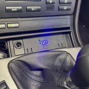 May include: Interior car detail showing a black dashboard with climate controls, a gear shift, and a digital display reading "12.7". The display is illuminated with a blue light. The gear shift is black leather.