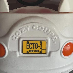 Puede incluir: Un coche de juguete de pl&aacute;stico blanco con las palabras COZY COUPE grabadas en la parte trasera. Cuenta con una matr&iacute;cula amarilla con el texto negro ECTO-1 y NEW YORK, flanqueada por dos luces circulares de pl&aacute;stico naranja a los lados.