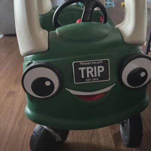 Puede incluir: Un coche de juguete de pl&aacute;stico verde con grandes pilares blancos, un volante negro y dos ojos redondos. El frente presenta una matr&iacute;cula negra con texto blanco que dice ESSEX FELLS TRIP EST 2024 y una boca roja curva y ancha.