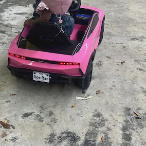 Puede incluir: Un coche de juguete el&eacute;ctrico de color rosa brillante con ruedas y molduras negras. La parte trasera cuenta con luces rojas, un aler&oacute;n negro y una matr&iacute;cula blanca que dice Xoe Rae Texas Est. 2024. El veh&iacute;culo tiene un asiento negro.