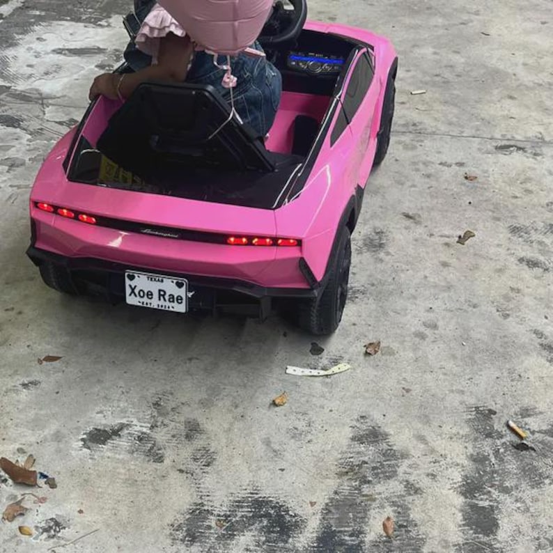 Puede incluir: Un coche de juguete el&eacute;ctrico de color rosa brillante con ruedas y molduras negras. La parte trasera cuenta con luces rojas, un aler&oacute;n negro y una matr&iacute;cula blanca que dice Xoe Rae Texas Est. 2024. El veh&iacute;culo tiene un asiento negro.