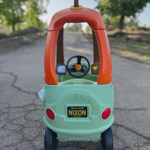 Puede incluir: Un coche de juguete de pl&aacute;stico verde claro y naranja con un volante negro en el interior. La parte trasera tiene una matr&iacute;cula negra con el texto NIXON, CALIFORNIA, EST 2023 y las palabras COZY COUPE grabadas en el cuerpo de pl&aacute;stico.