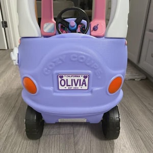 Puede incluir: Un coche de juguete de pl&aacute;stico color lavanda con pilares de techo blancos, volante negro y faros circulares naranjas. El frente presenta un logo 'COZY COUPE' moldeado y una matr&iacute;cula personalizada que dice 'CALIFORNIA OLIVIA EST 2024'.
