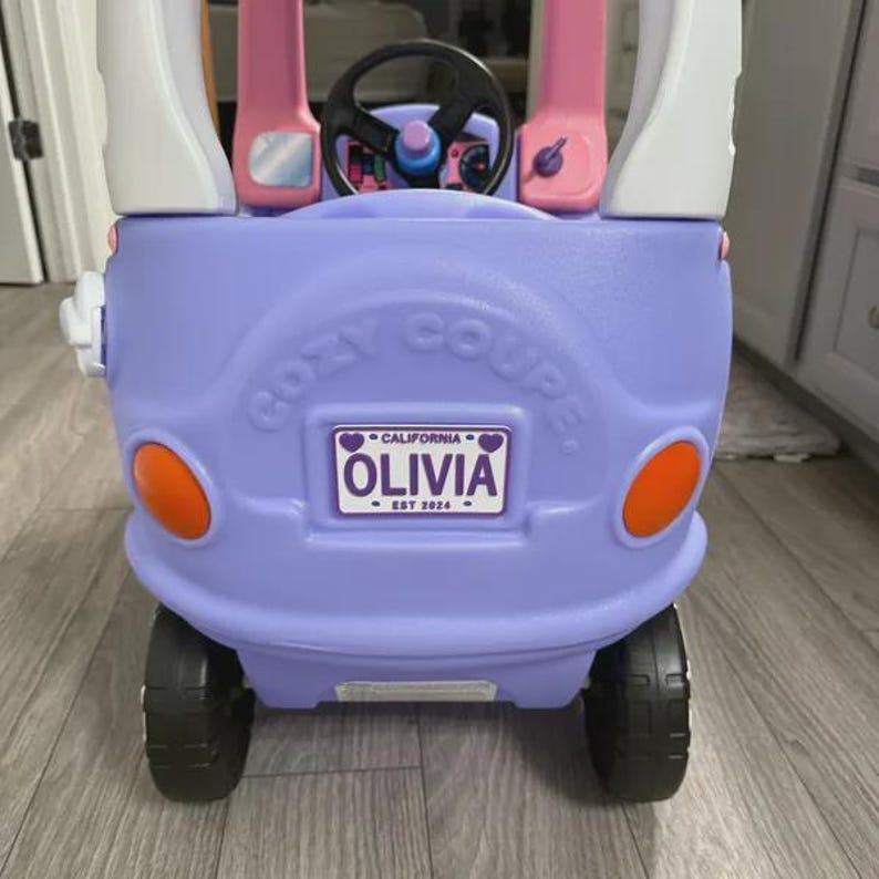 Puede incluir: Un coche de juguete de pl&aacute;stico color lavanda con pilares de techo blancos, volante negro y faros circulares naranjas. El frente presenta un logo 'COZY COUPE' moldeado y una matr&iacute;cula personalizada que dice 'CALIFORNIA OLIVIA EST 2024'.