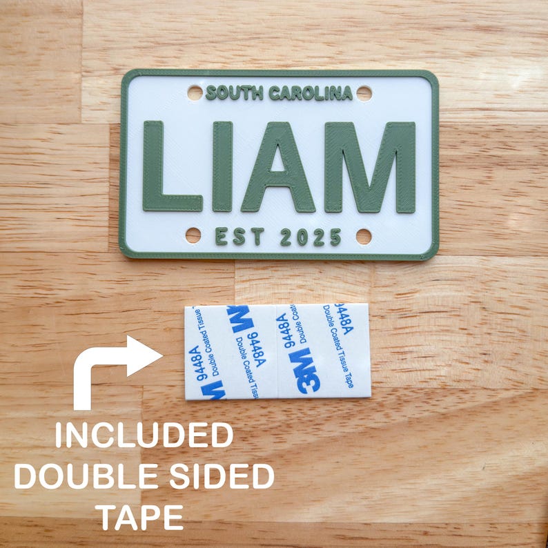 Puede incluir: Una placa de matr&iacute;cula rectangular blanca con borde verde y letras verdes que dicen SOUTH CAROLINA, LIAM y EST 2025. Debajo de la placa hay un trozo rectangular de cinta adhesiva de doble cara 3M 9448A con reverso blanco y texto azul.