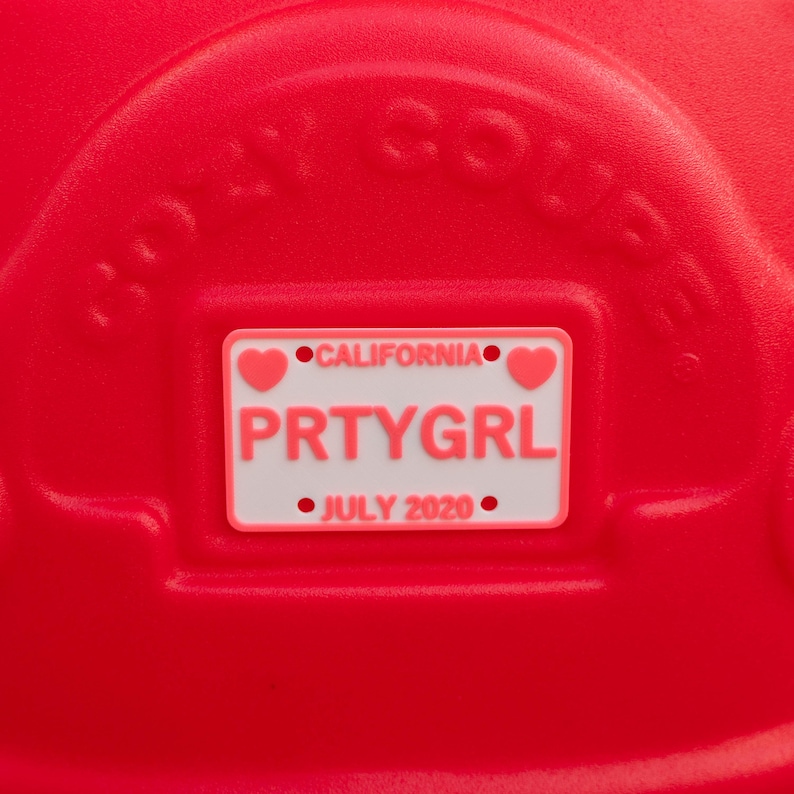 Puede incluir: Una superficie de pl&aacute;stico rojo brillante con el texto en relieve COZY COUPE y una placa de matr&iacute;cula rectangular blanca. La placa muestra el texto CALIFORNIA, PRTYGRL y JULY 2020, acentuado con dos peque&ntilde;os corazones rosas a los lados.
