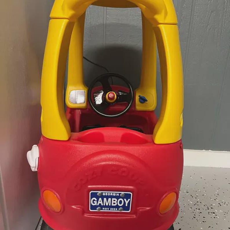 Puede incluir: Un coche de juguete de pl&aacute;stico rojo y amarillo con techo, volante negro y una matr&iacute;cula azul que dice GEORGIA GAMBOY NOV 2024. La parte delantera del veh&iacute;culo presenta faros circulares naranjas y una tapa de combustible blanca en el lateral.