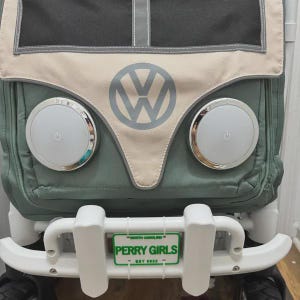 Puede incluir: Un cochecito de tela verde salvia y beige con un logotipo de Volkswagen, dos luces blancas circulares con s&iacute;mbolos de encendido y un parachoques delantero blanco. Una peque&ntilde;a matr&iacute;cula verde y blanca dice NORTH CAROLINA PERRY GIRLS EST 2022.