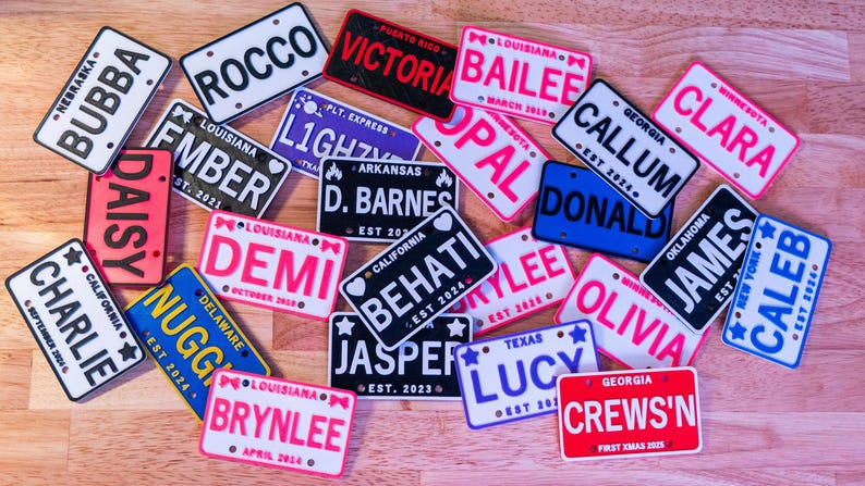 Puede incluir: Una colecci&oacute;n de placas de matr&iacute;cula en miniatura rectangulares y personalizadas en varios colores, cada una con el nombre de un estado, un nombre y una fecha. Las placas muestran nombres como Bubba, Rocco, Victoria, Bailee, Opal, Callum, Clara, Ember y otros.