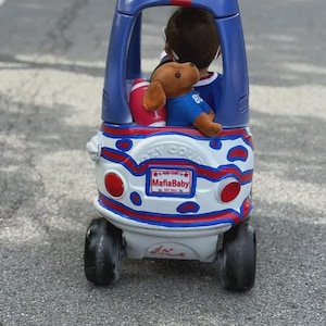 Puede incluir: Un coche de juguete azul y blanco con detalles rojos, que presenta una matr&iacute;cula con el texto New York MafiaBaby Est 2011. Dentro del coche hay un perro de peluche marr&oacute;n con una camiseta azul y un bal&oacute;n de f&uacute;tbol americano rojo y blanco.
