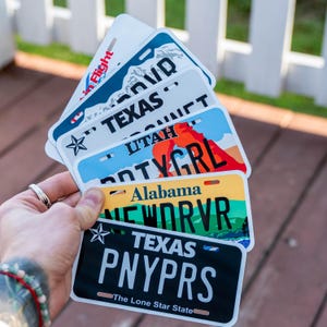 May include: A collection of license plates featuring various state designs. The plates display the names of states like Texas, Utah, and Alabama, with colorful graphics and unique license plate numbers. The plates are held in a hand.