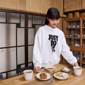 May include: A woman wearing a white sweatshirt with the text "JUST DO IT" in black dripping letters. She is standing in a kitchen setting and holding a plate of food. There are other plates of food on a wooden table.