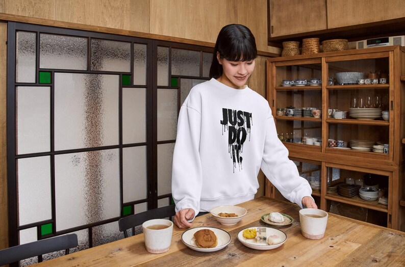 May include: A woman wearing a white sweatshirt with the text "JUST DO IT" in black dripping letters. She is standing in a kitchen setting and holding a plate of food. There are other plates of food on a wooden table.