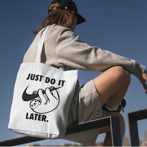 May include: White tote bag with black text and a sloth graphic. The bag reads "JUST DO IT LATER." The sloth is hanging from a Nike swoosh. The bag has white straps and is being carried by a person.