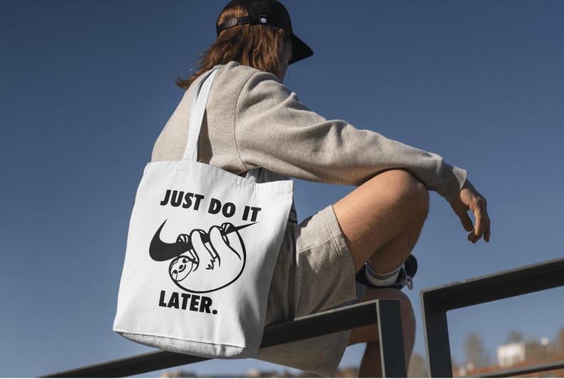 May include: White tote bag with black text and a sloth graphic. The bag reads "JUST DO IT LATER." The sloth is hanging from a Nike swoosh. The bag has white straps and is being carried by a person.