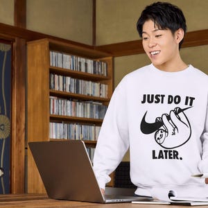 May include: White sweatshirt with the text "JUST DO IT LATER" and a sloth graphic. The sweatshirt features a black Nike swoosh incorporated into the sloth design. The person is smiling and holding a pen and a laptop.