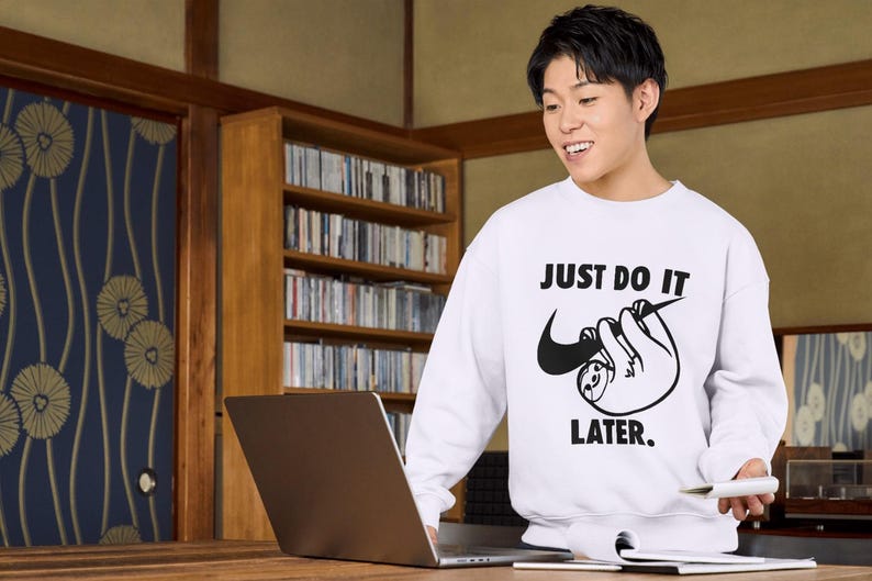 May include: White sweatshirt with the text "JUST DO IT LATER" and a sloth graphic. The sweatshirt features a black Nike swoosh incorporated into the sloth design. The person is smiling and holding a pen and a laptop.