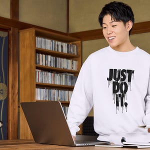May include: A white sweatshirt with black text that reads "JUST DO IT." The text is dripping down the sweatshirt. A person is wearing the sweatshirt and is looking at a laptop.