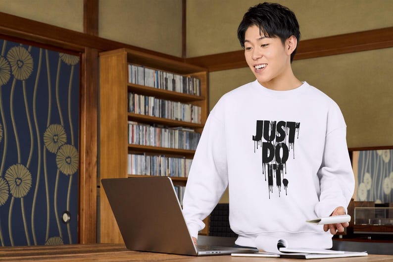 May include: A white sweatshirt with black text that reads "JUST DO IT." The text is dripping down the sweatshirt. A person is wearing the sweatshirt and is looking at a laptop.