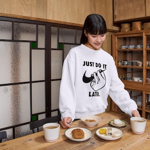 May include: White sweatshirt with the text "JUST DO IT LATER" and a sloth graphic. The image shows a wooden table with plates of food and cups. A wooden cabinet with dishes is in the background.
