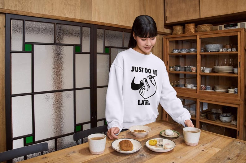May include: White sweatshirt with the text "JUST DO IT LATER" and a sloth graphic. The image shows a wooden table with plates of food and cups. A wooden cabinet with dishes is in the background.
