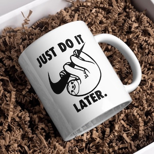 May include: White ceramic mug with a black sloth graphic and the text "JUST DO IT. LATER." The mug is inside a white box filled with brown crinkle paper, ideal for gifting.